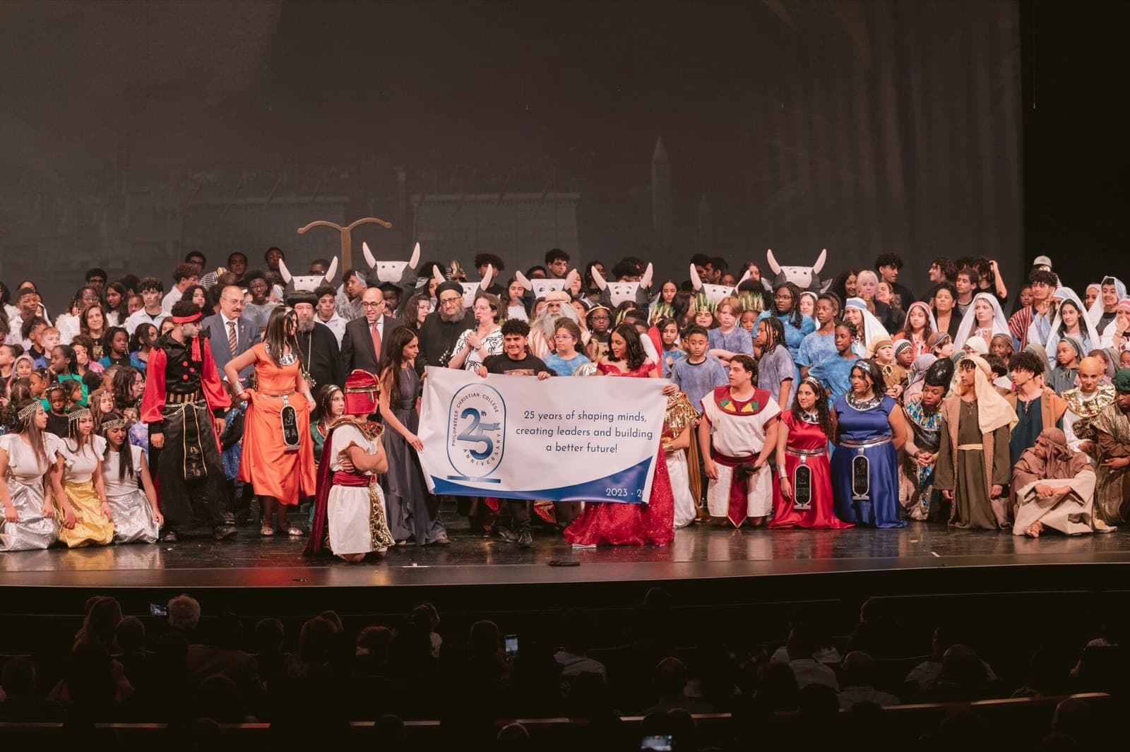 The whole cast of a school production stands on stage in costume, holding a banner that reads 25 years of shaping minds, creating leaders and building a better future.