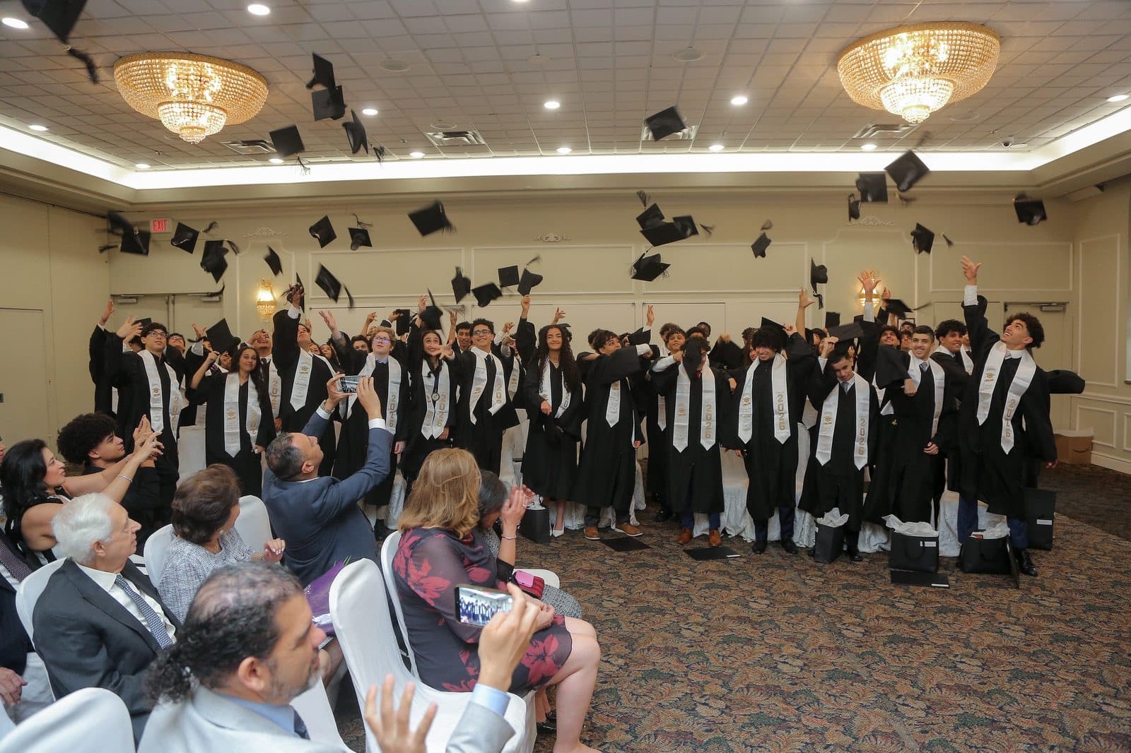 Graduates in black gowns and white stoles toss their caps into the air in a banquet hall while family and friends watch from their seats.