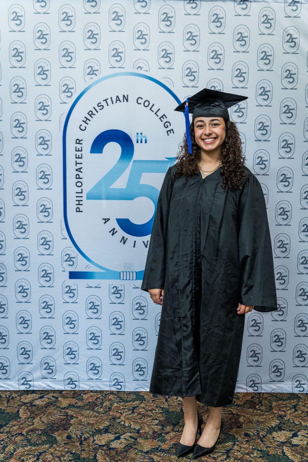 A graduate in cap and gown smiles in front of the school's 25th anniversary backdrop.