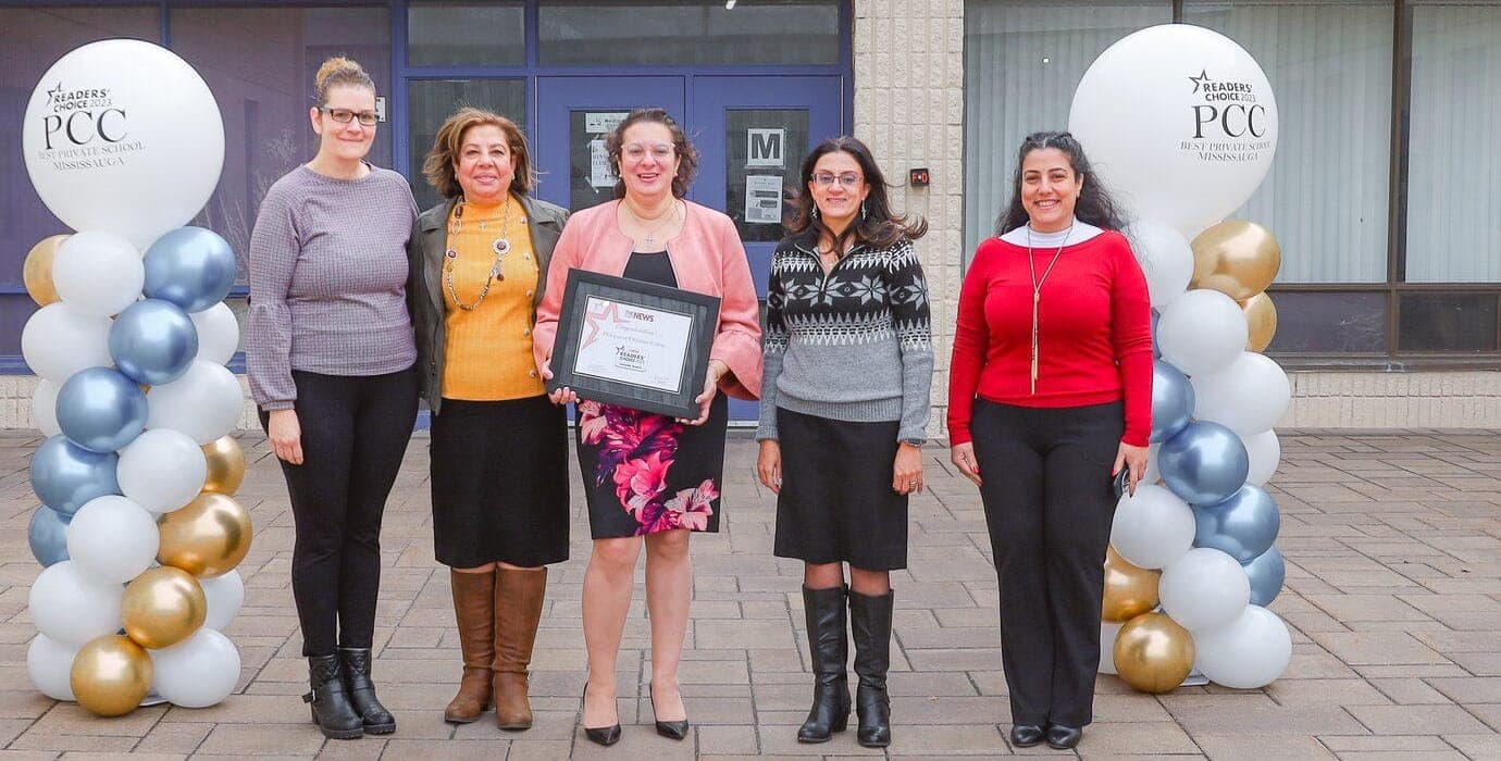 Five members of the PCC staff stand together in front of the school entrance and its sign, between two columns of silver, white and gold balloons, one of them holding a framed award.